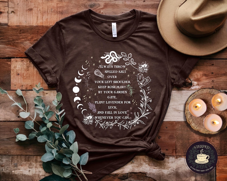 May include: A brown t-shirt with a white graphic design featuring a moon phase, a snake, and text that reads "Always throw spilled salt over your left shoulder. Keep rosemary by your garden gate, plant lavender for luck, and fall in love whenever you can."