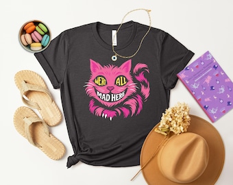 チェシャ猫と「We're All Mad Here」のトレンドグラフィックTシャツ。ピンク、ブルー、パープルのカラーバリエーション。『不思議の国のアリス』ファンや紅茶好きの方へのギフトに最適です。