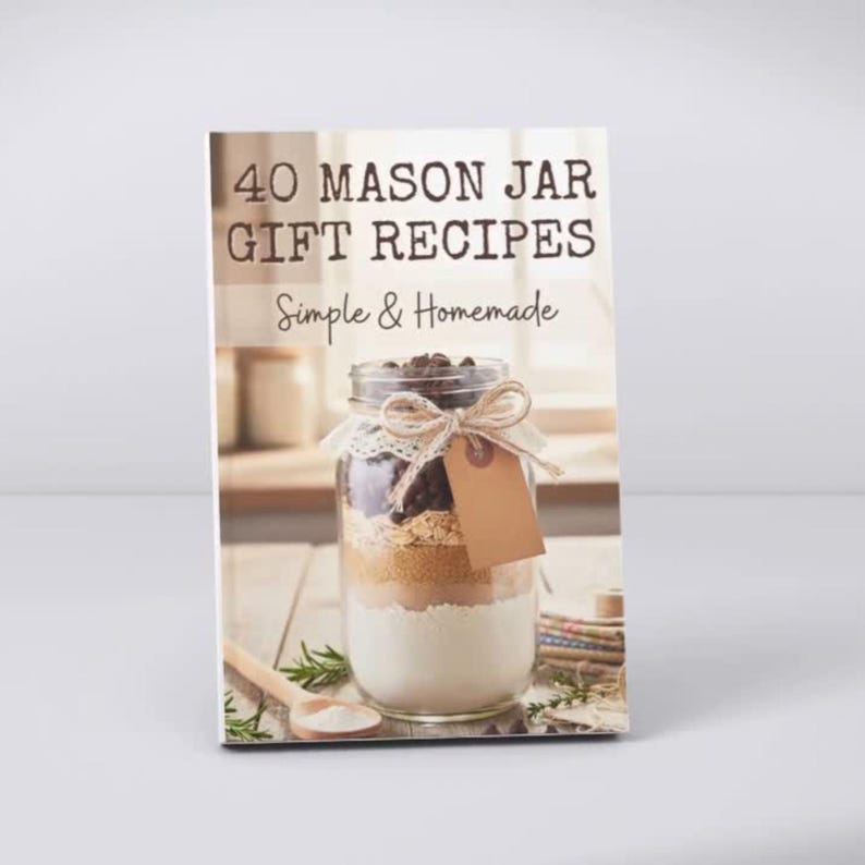 May include: A book titled 40 MASON JAR GIFT RECIPES Simple & Homemade featuring a glass jar filled with layered dry ingredients, tied with a twine bow and a blank brown paper tag, resting on a wooden surface with a wooden spoon nearby.
