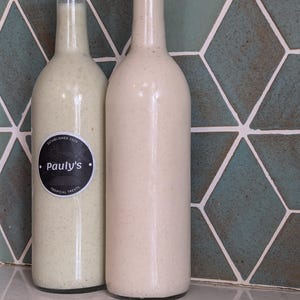May include: Two glass bottles with white liquid and black caps. The bottle on the left has a black and white label that says "Established 2023 Pauly's Tropical Treats".