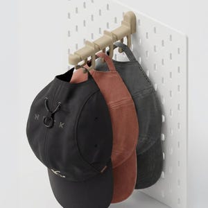 Puede incluir: Un panel perforado blanco con un sistema de ganchos beige que sostiene cuatro gorras de béisbol. Las gorras son negras, óxido y grises. La gorra negra tiene las letras "H K" en la parte delantera. El panel tiene agujeros espaciados.