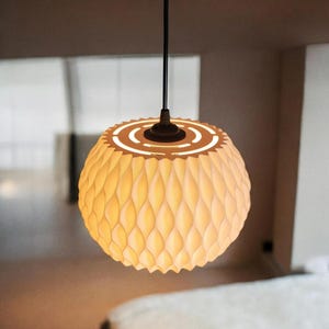Peut inclure: Suspension luminaire sphérique avec abat-jour texturé. L'abat-jour présente un motif en losanges répétés et émet une lueur jaune chaude. La lumière est suspendue à un cordon noir et possède un dessus marron foncé. Diamètre d'environ 30 cm.