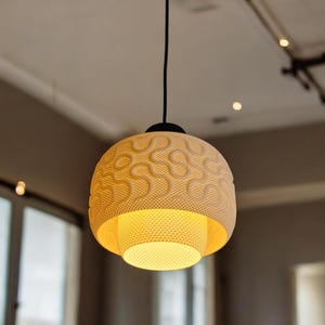 May include: A cream-coloured, spherical pendant light fitting with a patterned exterior and a textured, open bottom. The light hangs from a black cord and emits a warm, yellow glow. The fitting is a modern home decor item.