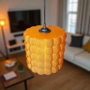 Peut inclure: Suspension luminaire cylindrique jaune-orangé vif, avec un dessus festonné et un design texturé en couches. La lampe est suspendue à un cordon et un luminaire noirs. L'éclairage est dans un salon.
