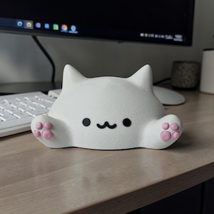 Op de afbeelding: Een wit katvormig beeldje met een getextureerd oppervlak. Het heeft zwarte ogen en een lachende mond. De poten zijn roze. De kat zit op een houten bureau.