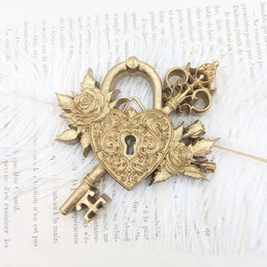 Peut inclure: Un cadenas doré en forme de cœur avec une clé ornée insérée dans la serrure. Le cadenas présente des volutes complexes et est entouré de roses et de feuilles sculptées de chaque côté, formant un objet décoratif en métal détaillé.