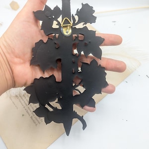 May include: A black metal wall decoration in a floral design. The back has a gold-coloured hanger. The piece is held in a hand against a white background with a vintage book page.