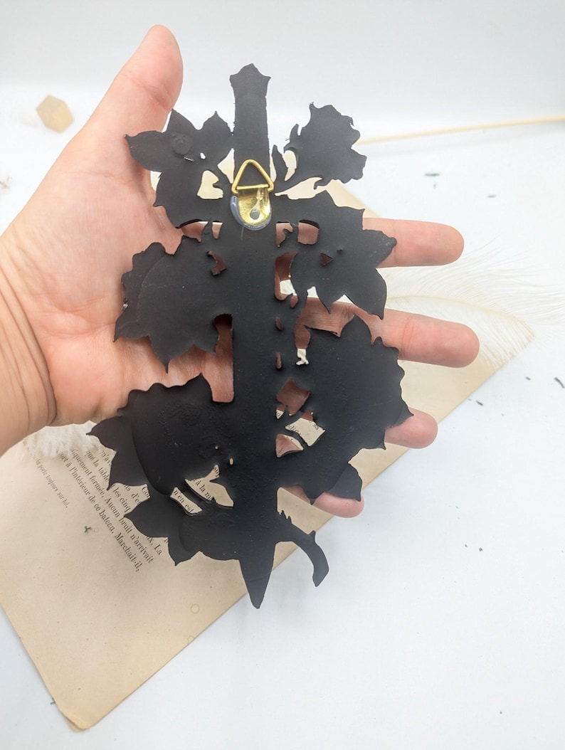 May include: A black metal wall decoration in a floral design. The back has a gold-coloured hanger. The piece is held in a hand against a white background with a vintage book page.
