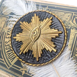May include: A gold and black circular pendant with a stylized eye design in the center. The eye is surrounded by a radiating sunburst pattern.