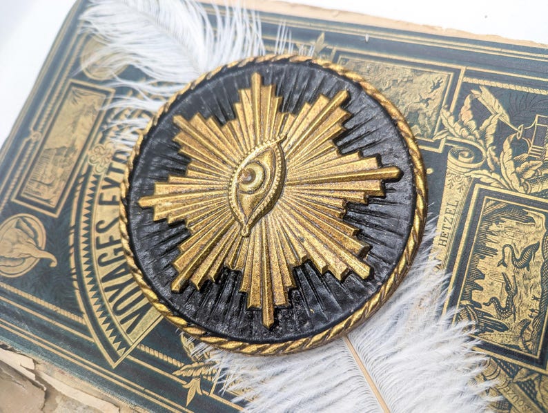 May include: A gold and black circular pendant with a stylized eye design in the center. The eye is surrounded by a radiating sunburst pattern.