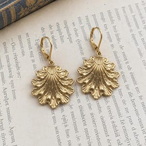 Könnte beinhalten: Ein Paar goldfarbene Ohrhänger mit einem detaillierten, fächerförmigen Muscheldesign, das strahlenförmige Rillen und gewellte Kanten aufweist. Jeder Ohrring ist an einem sicheren Klappbügel befestigt und besitzt eine metallische Oberfläche.