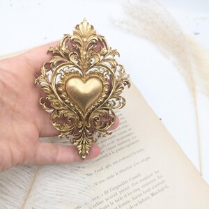 May include: Ornate gold-colored metal heart-shaped brooch with intricate scrollwork and a central heart design. The brooch is a vintage-style accessory, likely for clothing or decorative purposes.
