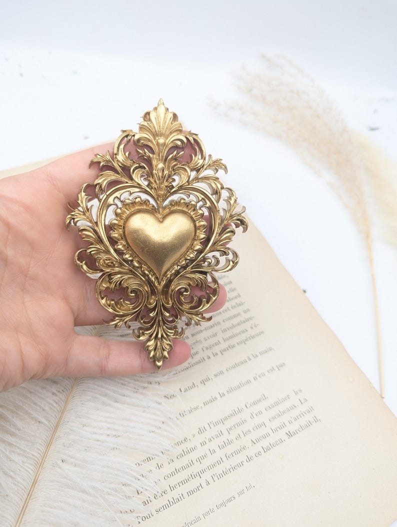 May include: Ornate gold-colored metal heart-shaped brooch with intricate scrollwork and a central heart design. The brooch is a vintage-style accessory, likely for clothing or decorative purposes.