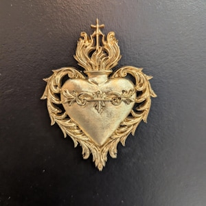 May include: A gold-colored wall ornament shaped like a Sacred Heart, featuring intricate scrollwork and acanthus leaf detailing surrounding the central heart. A small cross sits atop a flame motif at the peak of the heart-shaped decorative piece.