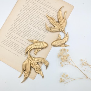 May include: Gold-colored metal koi fish sculptures. The fish are detailed with fins and scales, and are arranged on a vintage book page. Dried flowers are in the background. The sculptures are decorative.