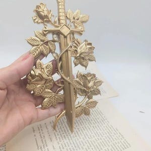 May include: A gold-coloured decorative sword featuring rose and leaf detailing. The sword has a cross-shaped hilt and is surrounded by detailed floral elements. The design is intricate and ornate, with a vintage aesthetic.