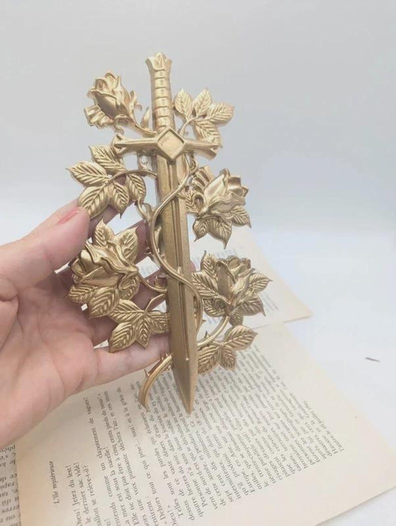 May include: A gold-coloured decorative sword featuring rose and leaf detailing. The sword has a cross-shaped hilt and is surrounded by detailed floral elements. The design is intricate and ornate, with a vintage aesthetic.