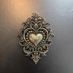 May include: Ornate, antique-style heart-shaped wall decor. The heart is centered within a decorative frame of swirling, leaf-like patterns. The piece has a metallic gold finish, set against a dark background.