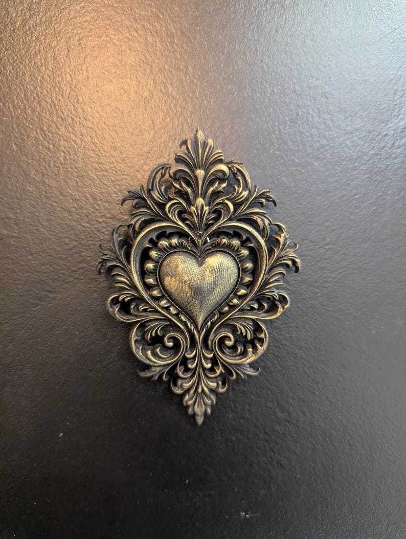 May include: Ornate, antique-style heart-shaped wall decor. The heart is centered within a decorative frame of swirling, leaf-like patterns. The piece has a metallic gold finish, set against a dark background.