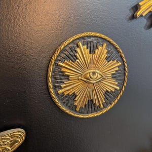 May include: A gold-colored eye with rays emanating from it, set against a black background. The eye is surrounded by a gold rope-like border.