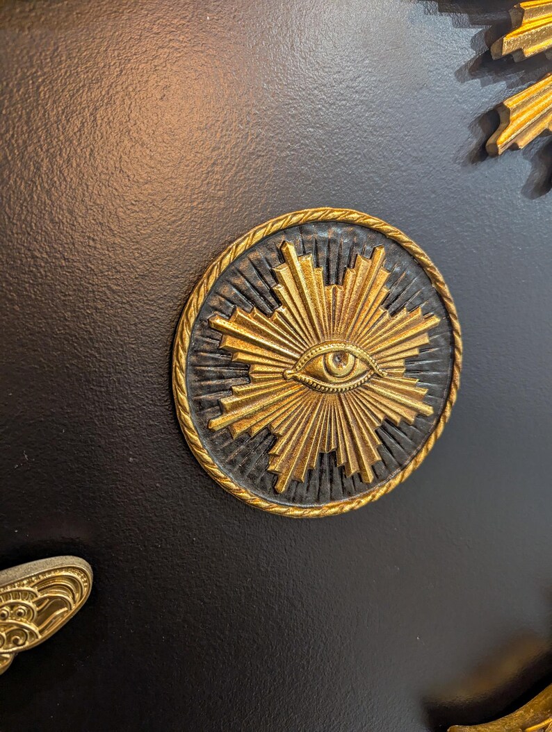May include: A gold-colored eye with rays emanating from it, set against a black background. The eye is surrounded by a gold rope-like border.