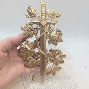 May include: A gold-coloured decorative sword with a floral design. The sword is surrounded by roses and leaves, creating an elegant and artistic piece. The sword is held in a hand.