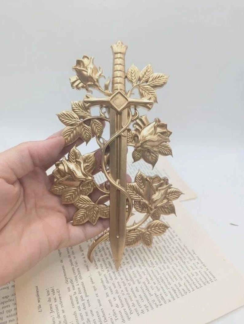 May include: A gold-coloured decorative sword with a floral design. The sword is surrounded by roses and leaves, creating an elegant and artistic piece. The sword is held in a hand.