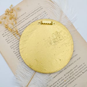 May include: A gold-colored round wall decor with a textured surface. The decor has a metal hanger on the back.