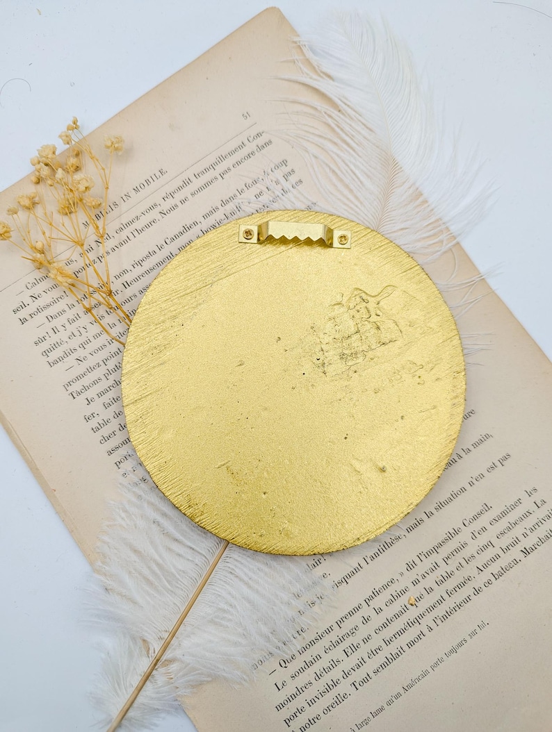 May include: A gold-colored round wall decor with a textured surface. The decor has a metal hanger on the back.