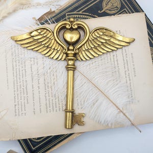 May include: A gold-colored decorative key featuring a central heart shape flanked by two detailed wings extending horizontally. The key has a long, slender shaft and a standard bit at the bottom, crafted with a metallic finish and ornate relief details.
