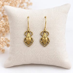 Può includere: Un paio di orecchini pendenti in metallo color oro con un design dettagliato a forma di scarabeo. Ogni orecchino è composto da un ciondolo a forma di scarabeo scolpito sospeso a un semplice gancio curvo, con finitura metallica testurizzata.