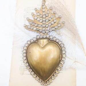 Può includere: Medaglione vintage a forma di cuore in ottone, con una parte superiore a forma di corona ornata di cristalli trasparenti. Il medaglione è aperto, rivelando un interno vuoto. Poggia su una pagina di libro vintage con una piuma ed erba secca.