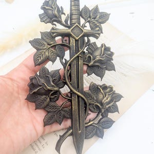 May include: Bronze decorative sword with roses and leaves. The sword has a cross-shaped hilt and a crown-like pommel. Roses and leaves are entwined around the sword's blade, creating an elegant, gothic-inspired design.