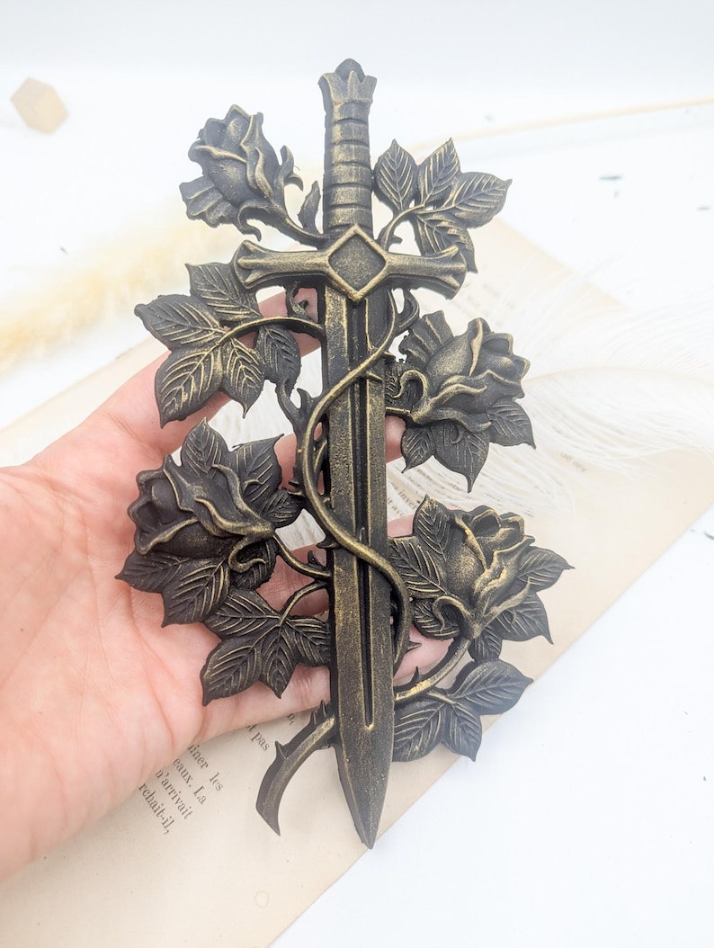 May include: Bronze decorative sword with roses and leaves. The sword has a cross-shaped hilt and a crown-like pommel. Roses and leaves are entwined around the sword's blade, creating an elegant, gothic-inspired design.