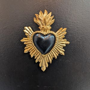 May include: A decorative wall ornament featuring a central smooth black heart shape surrounded by a gold-colored metal frame with radiating sunburst rays and a flame-like topper. The piece has a detailed beaded border around the heart.