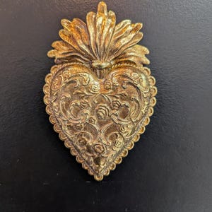 May include: A gold-colored heart-shaped wall ornament featuring an intricate embossed floral and scrollwork pattern. The piece is topped with a stylized flame design and has a scalloped edge, showcasing a metallic finish with dark highlights.