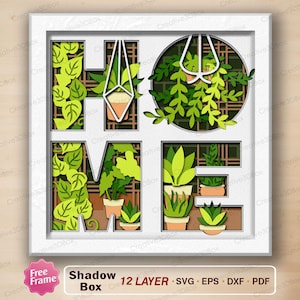 Puede incluir: Una caja de sombra enmarcada en blanco con la palabra "HOME" recortada, llena de plantas de papel en capas en tonos verdes y marrones. El diseño incluye plantas colgantes y suculentas en macetas. El texto "Shadow Box" y "12 LAYER" son visibles.