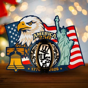 K&ouml;nnte beinhalten: Patriotische Holzdekoration mit einem Adler, der amerikanischen Flagge, der Freiheitsstatue und einer Laterne mit dem Text "US" und "America 250". Das Design umfasst die Liberty Bell und die Jahre 1776 und 2026, in Rot, Wei&szlig;, Blau und Gold.