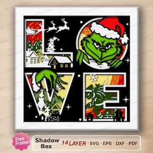 Pode incluir: Uma obra de arte em caixa de sombra em camadas com a palavra "LOVE" formada por recortes. O design apresenta o Grinch usando um chapéu de Papai Noel, imagens de Natal e uma cena de vila nevada. A obra de arte é emoldurada em branco.