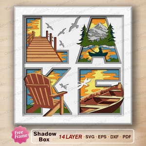May include: A square multi-layered shadow box art piece featuring the word LAKE. Each letter contains a scene: L shows a wooden dock, A shows a mountain with a person in a canoe, K shows an Adirondack chair, and E shows a rowboat on water.