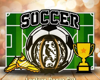 May include: A paper cut design featuring a soccer theme. The design includes a soccer ball, a trophy, soccer cleats, and the word "SOCCER". A lantern with a soccer player silhouette is in the center. The background is a green soccer field.