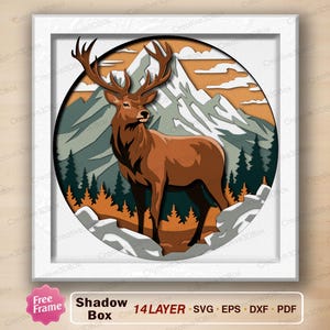 May include: A multi-layered shadow box art piece featuring a brown deer with large antlers standing in a mountain landscape. The circular design includes layered cutouts of snow-capped mountains, evergreen trees, and rocky terrain in shades of brown, green, and white, all enclosed within a square white frame.