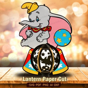Puede incluir: Un diseño recortado en papel con Dumbo el elefante, una pelota azul con estrellas amarillas y una linterna de circo. La linterna tiene un marco negro con recortes de estrellas y una imagen de un elefante. El texto "Lantern Paper Cut" está en la parte inferior.