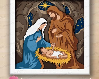 May include: A multi-layered paper shadow box art piece depicting a nativity scene with a figure in a blue robe and white head covering, a figure in a brown robe, and a baby in a manger, set against a dark cave background with a yellow star.
