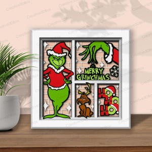 May include: A framed Christmas-themed artwork featuring the Grinch in a red Santa suit, a green hand, a reindeer, and the words "MERRY GRINCHMAS." The artwork is set against a brick-like background with a white frame.