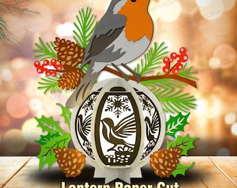 May include: A paper cut design featuring a gray and orange robin perched on a branch with pine needles, red berries, and a decorative lantern. The lantern has a bird and snowflake design. The words "Lantern Paper Cut" are at the bottom.