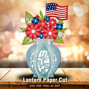 Op de afbeelding: Een lichtblauwe papieren lantaarn met een bloemstuk en een Amerikaanse vlag. De bloemen zijn rood, blauw en wit. De lantaarn heeft de tekst "US" en een hoedontwerp. De tekst "Lantern Paper Cut" staat onderaan.