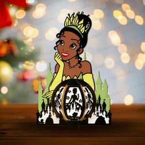 Puede incluir: Una linterna decorativa de papel con un diseño de princesa. La linterna tiene una base negra y un recorte circular con siluetas. La princesa está representada con un vestido verde y amarillo, con una corona y un castillo en el fondo.