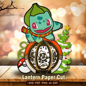 Könnte beinhalten: Eine Papierlaterne mit einem grünen Bulbasaur-Charakter mit einem rot-grünen Schal, einem Weihnachtsbaum und einem Pokéball. Die Laterne hat einen schwarzen Sockel und den Text "Lantern Paper Cut".