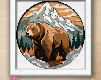 May include: A square white frame containing a circular layered paper art piece featuring a brown bear standing in the foreground. The background displays a mountain range with snow-capped peaks, dark green pine trees, and an orange sky with clouds.